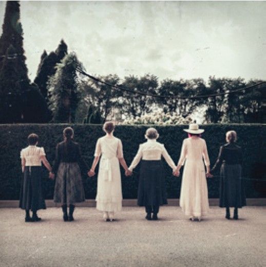Six women holding hands, viewed from behind, in front of a hedge and trees.