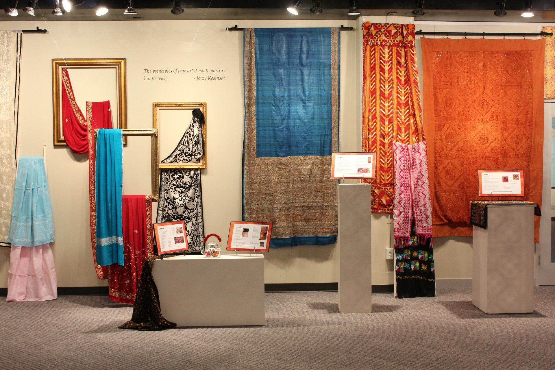 Museum display of various colorful textiles draped from picture frames and curtain rods