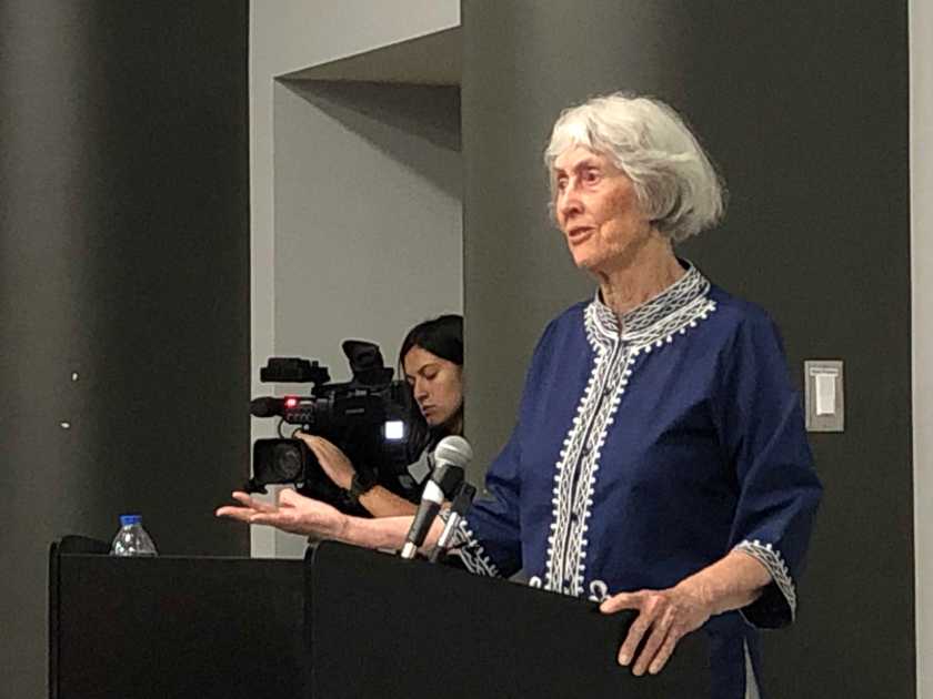 Older woman in navy embroidered tunic speaking at a podium, gesturing with left hand. Camerawoman filming in background.