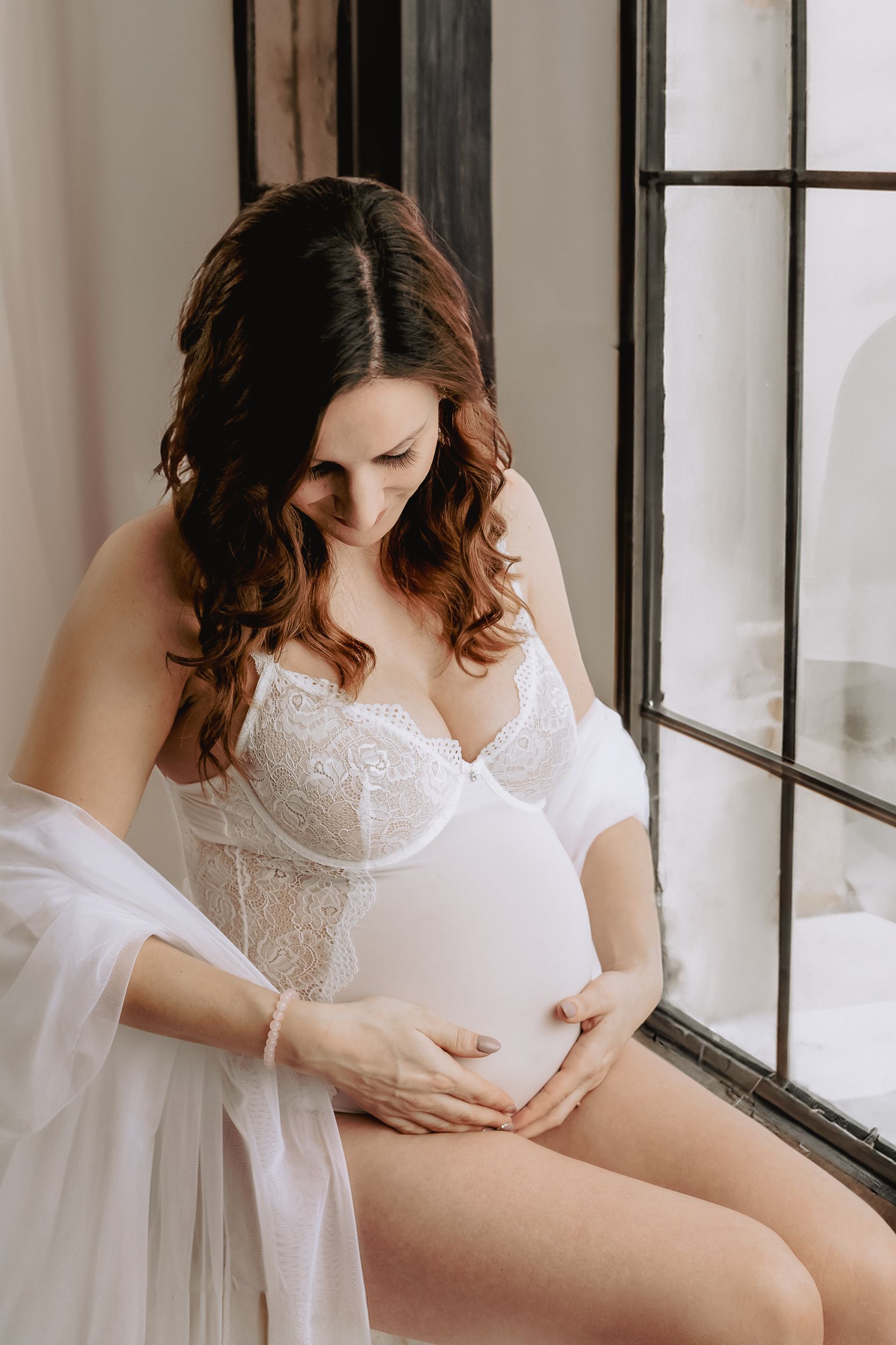 Eine schwangere Frau in Spitzenbody vor einem Fenster sitzend
