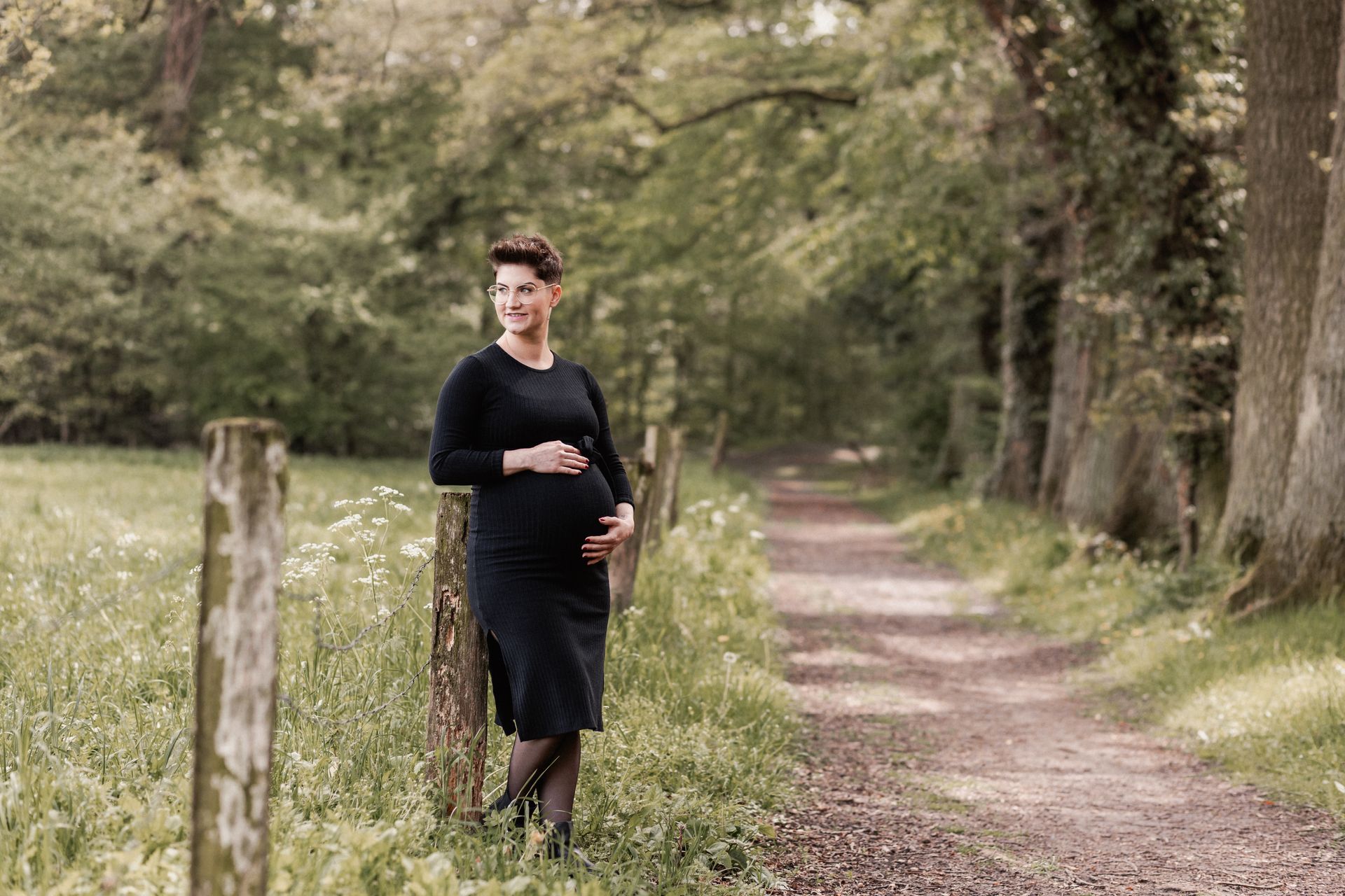 Schwangere in schwarzem Strickkleid am Zaunpfosten lehnend