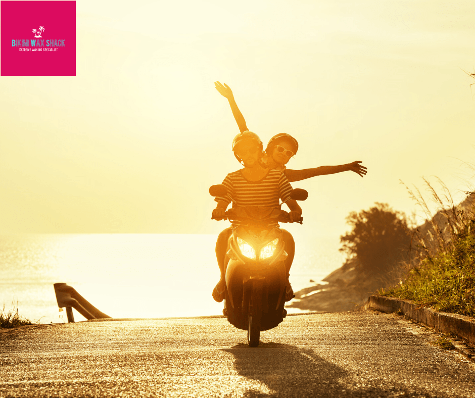 Male and female on a moped with woman's arms outstretched. Background is a sunset
