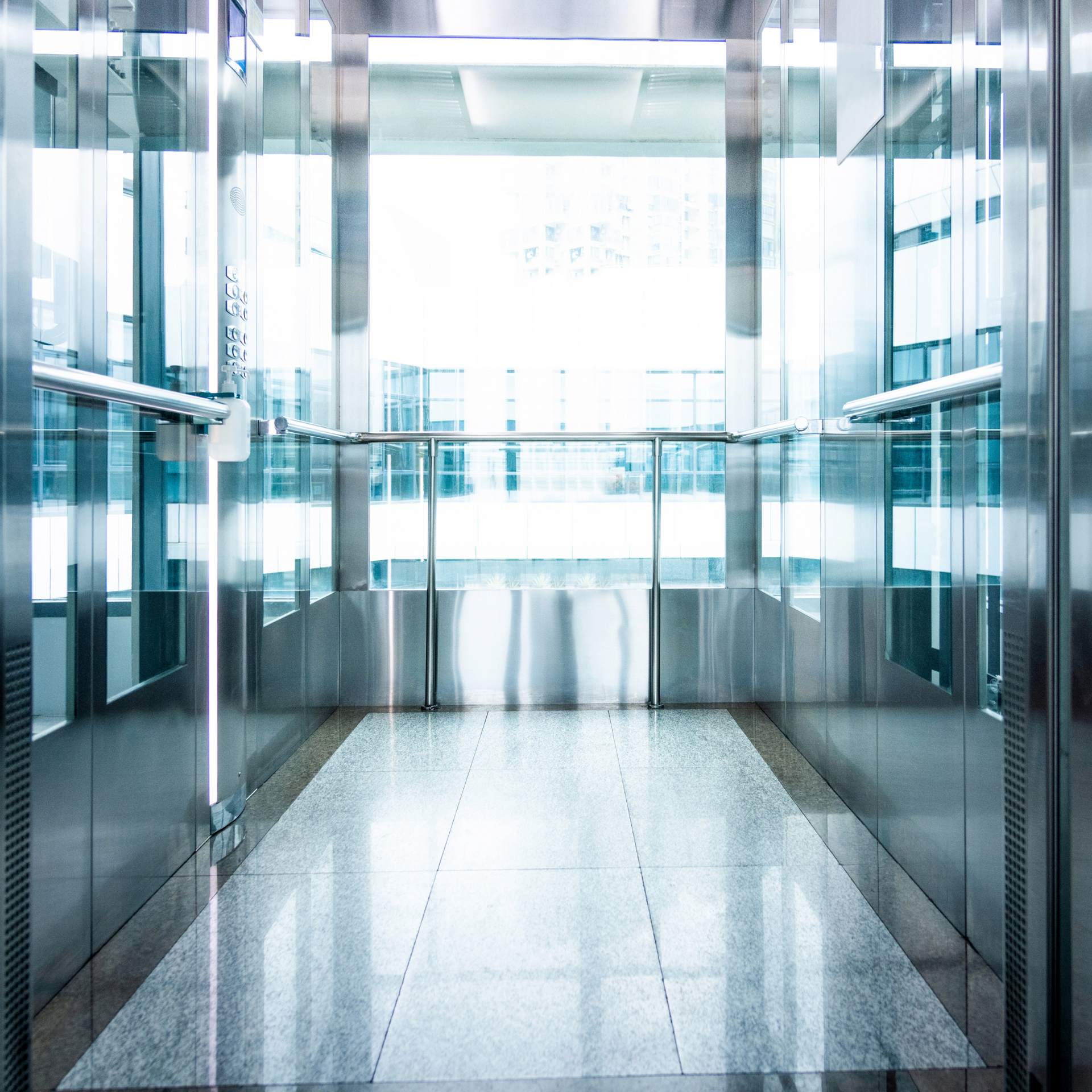 Ein leerer, lichtdurchfluteter Fahrstuhl mit Glaswänden und glänzenden Edelstahlflächen. Die klare,