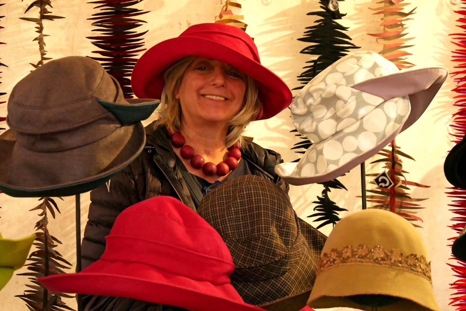 Angelika Klose inmitten ihrer Hüte, die sie kreiert hat - zwischen braun-dunkelgrünem Hut, das wie für die Jagd geeignet scheint, Regencape (weiße Kreise auf hellgrauem Grund und rosafarben auf der Rückseite bzw. Innenseite), rot, 20er Jahre-schick und Sherlock-Holmes-Imitat, lächelt sie zufrieden ins Publikum - im Hintergrund hängen die 