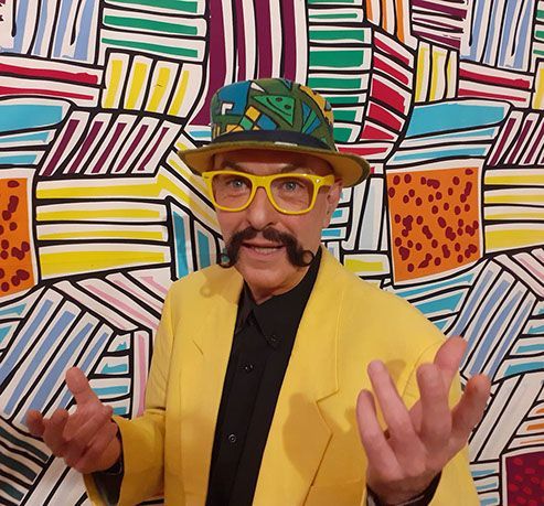 Carlos Klein zu sehen in seinem knallgelbem Jackett und mit seiner knallgelben Brille, der Pullover schwarz. Seine Hutkreation? Passend zum Outfit. Die Wand dahinter sowieso!