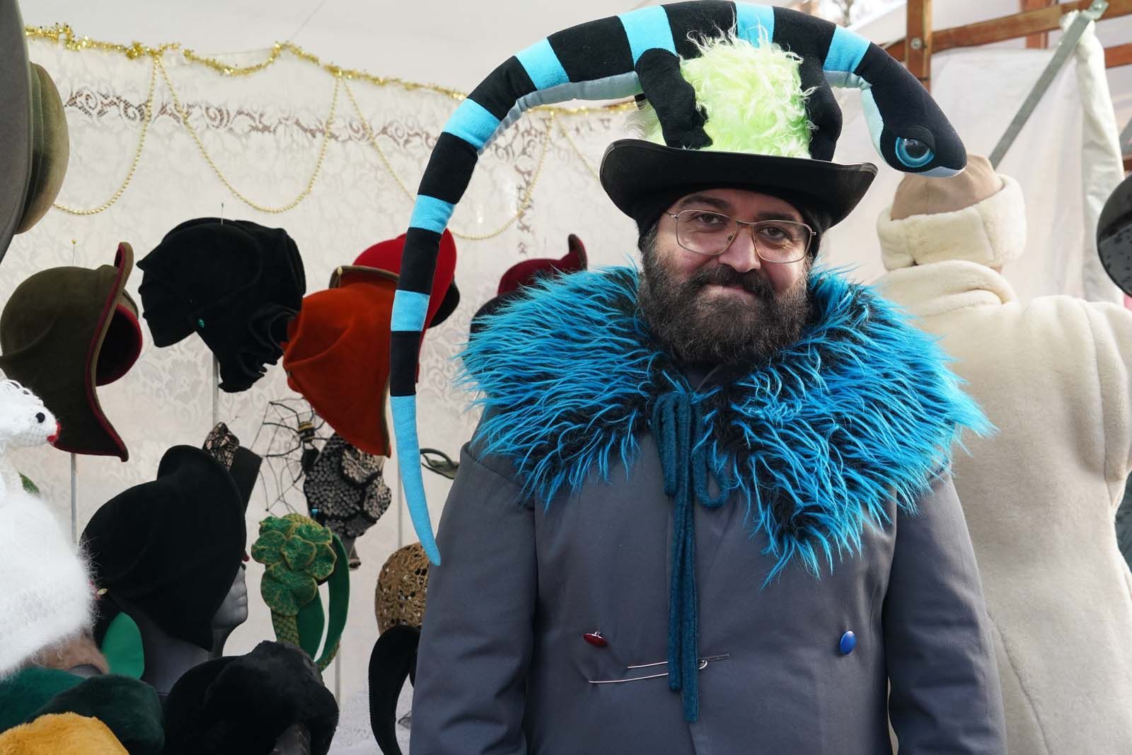 Gianni Klein ist Hatartist. Bei Weihnachten in Westend in Berlin-Charlottenburg steht er vor seinem Stand mit einer schwarz-blau geschwungenen Stoffschlange über dem Kopf. Gianni Klein ist bekannt für seine verspielten Hutkreationen.