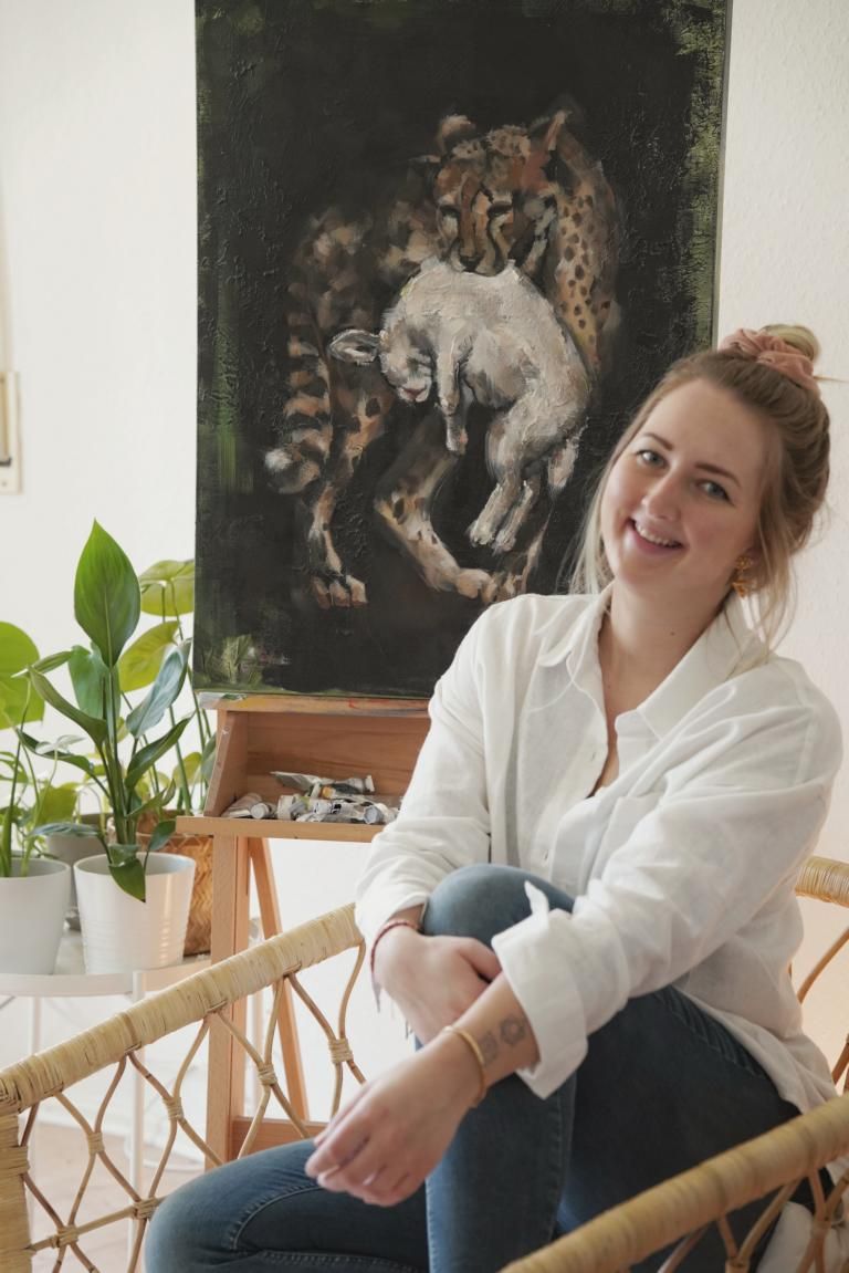 Jana Voscort ist eine junge Künstlerin in Berlin. Sie malt Bilder.  Auf dem Bild sitzt sie auf einem Korbstuhl, im Hintergrund an der Wand eins ihrer Bilder, das sie gemalt hat. Darauf ist ein totes Lamm zu sehen, das ein Leopard gerissen hat.