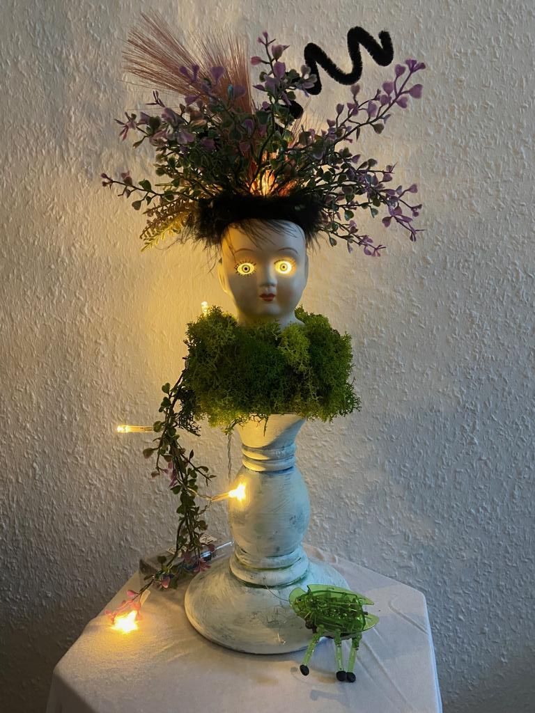 Großartig wirkt diese Lampe! Sie ist aus einem Puppenkopf, deren Augen leuchten, einer Vase als Körper und mit allerlei Moos um den Rumpf zusammengesetzt. Als Haare dienen ihr Trockenblumen, die ihr wild aus dem Kopf herauswachsen.