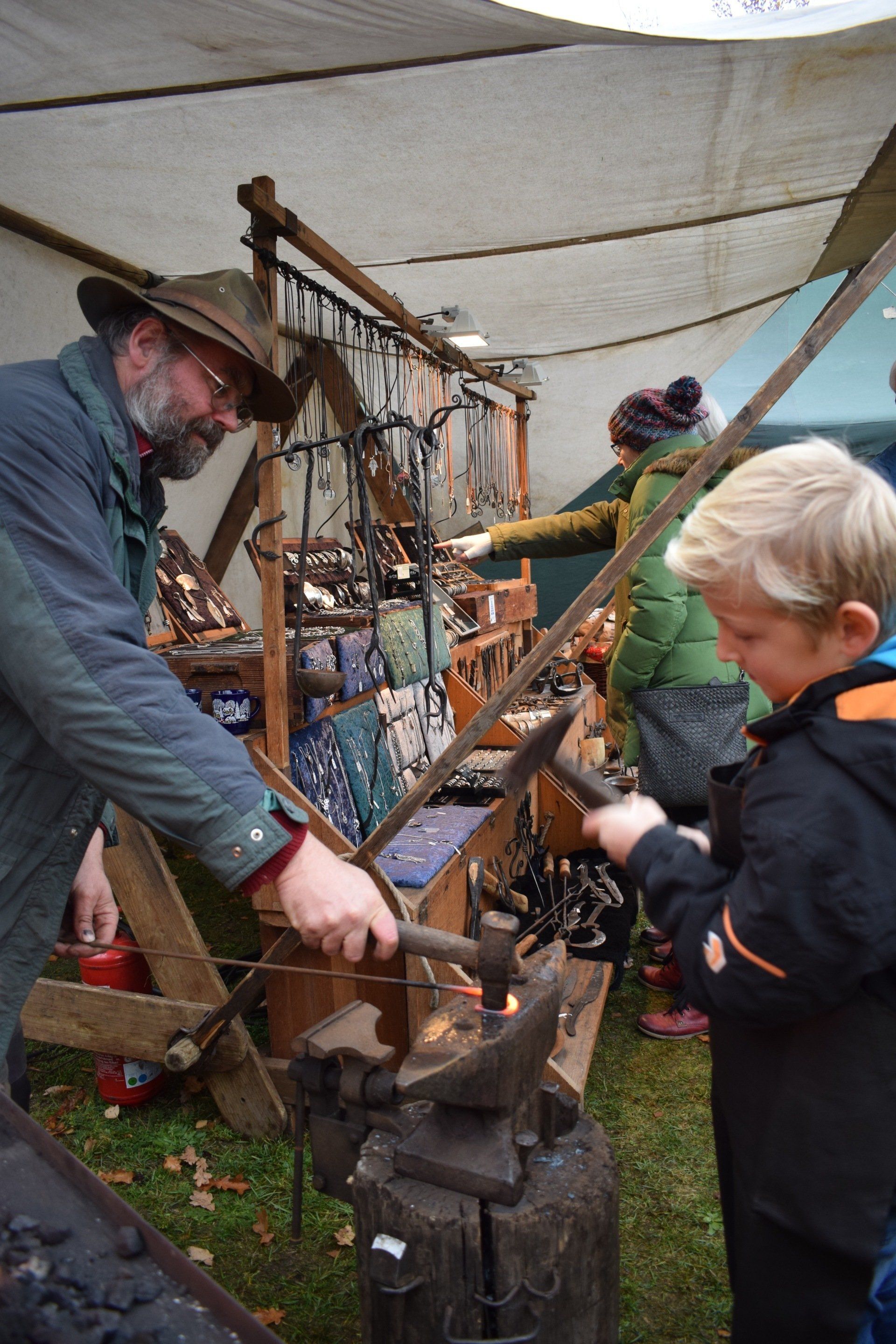 Schmied Falk Laxander an seinem Stand beim Wein- und Winzerfest Westend - Pfingsten in der Preußenallee. Er bearbeitet ein glühend heißes Werkstück mit Hand und hat sich die Zuarbeit eines etwa 10-jährigen blondhaarigen Jungen gesichert. An seinem Stand stellt Falk Laxander selbstgeschmiedete Kleinode wie Armreifen aus Silberbesteck, Ringe, Broschen, Haarspangen, Grillgabeln mit Widderkopf oder Griffen aus Langhorn vor.