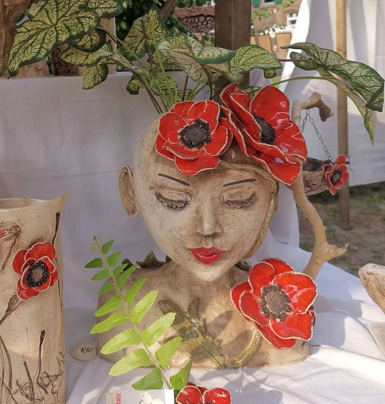 Martina Heinl bezeichnet diese Pflanztöpfe und Vasen als Mohnblütenkeramik. Mohnblüten kleben an der Vase und als Kopfschmuck an dem breiten, hübschen Gesicht eines Mädchens aus Keramik.