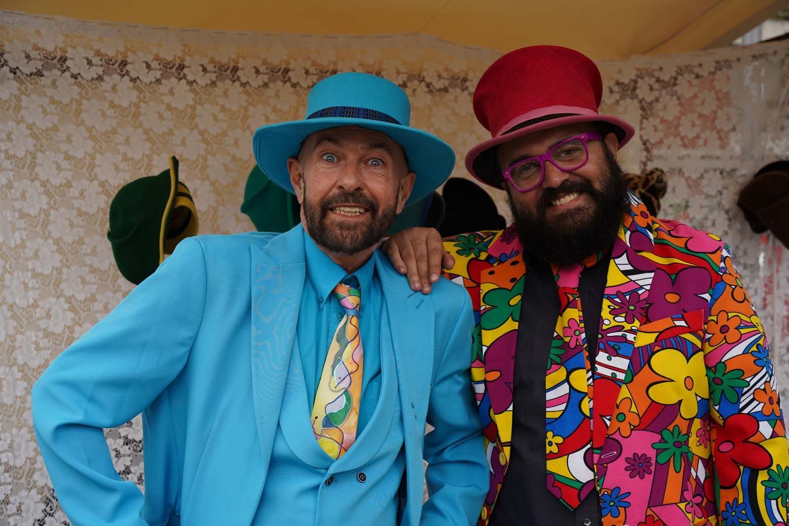Carlos und Gianni Klein haben die passende Kopfbedeckung und das passende Outfit für den Ostermarkt Zwei kunstvoll gekleidete Hutmacher in bunten Anzügen und auffälligen Hüten stehen vor einer Wand mit weiteren Hutmodellen