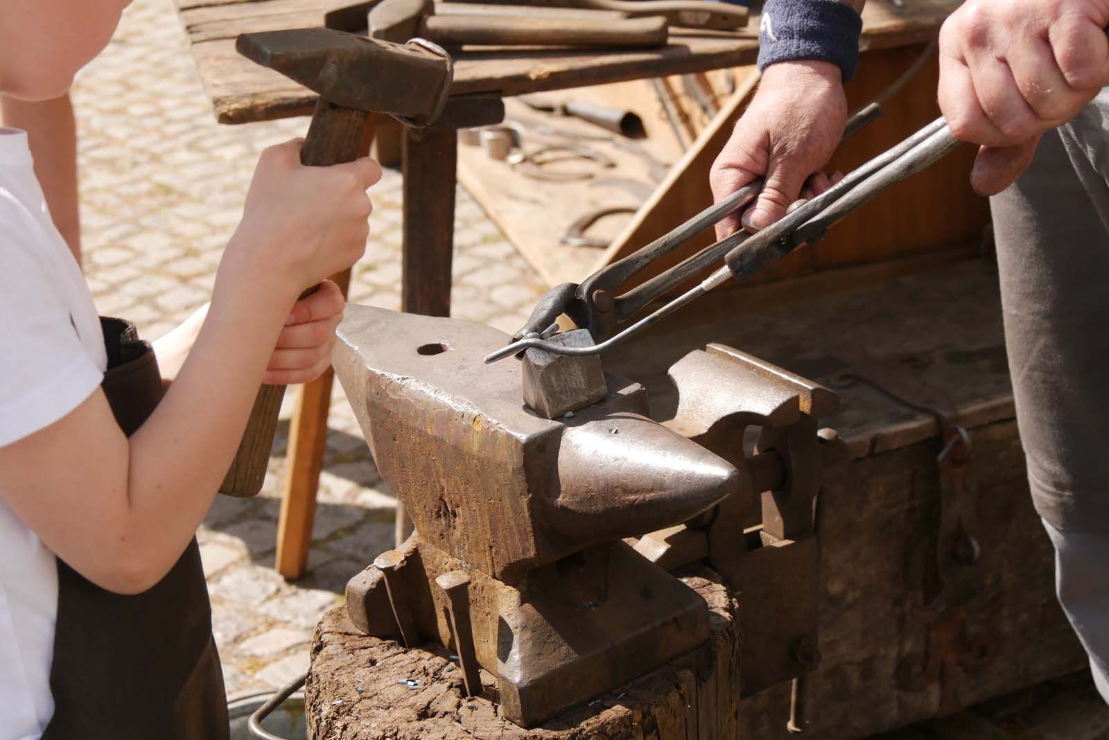 Ein Kind schmiedet mit dem Hammer auf dem Amboss, während Schmied Falk Laxander das Werkstück mit der Zange hält – Handwerk zum Mitmachen bei WeinPfingsten