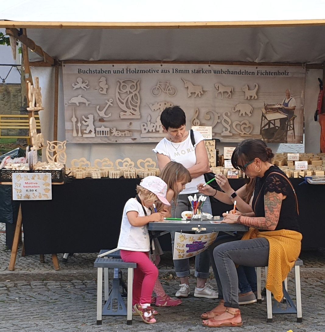 Basteltisch vor dem Stand von Holzhandwerker Jürgen Tschötschel – Kinder bemalen Buchstaben und Figuren aus Fichten- und Buchenholz
