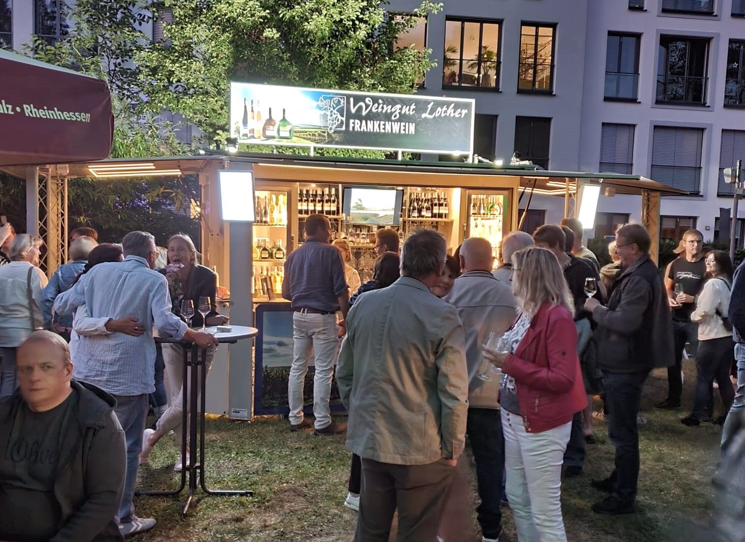 Weingut Lother aus Franken: Vor dem Weinstand der Winzerfamilie Lother stehen gutgekleidete Menschen bei einem wohligen Glas Wein. Es ist lauer Sommerabend. Das Licht aus dem Weinausschankwagen der Famlie lädt zum Verweilen ein.