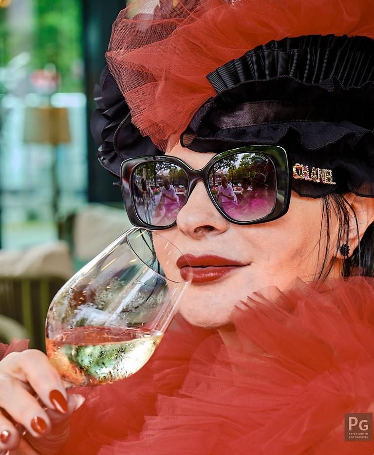 Jutta Seidenkranz vertritt Jutta Fashion in Charlottenburg-Wilmersdorf von Berlin. Sie hat ein knallrotes Federkleid als Jacke an und eine Kopfbedeckung aus schwarzen Federn mit rotem Tüll. Mit einem Glas Wein in der Hand spricht sie mit Künstlerin Silke Bach.