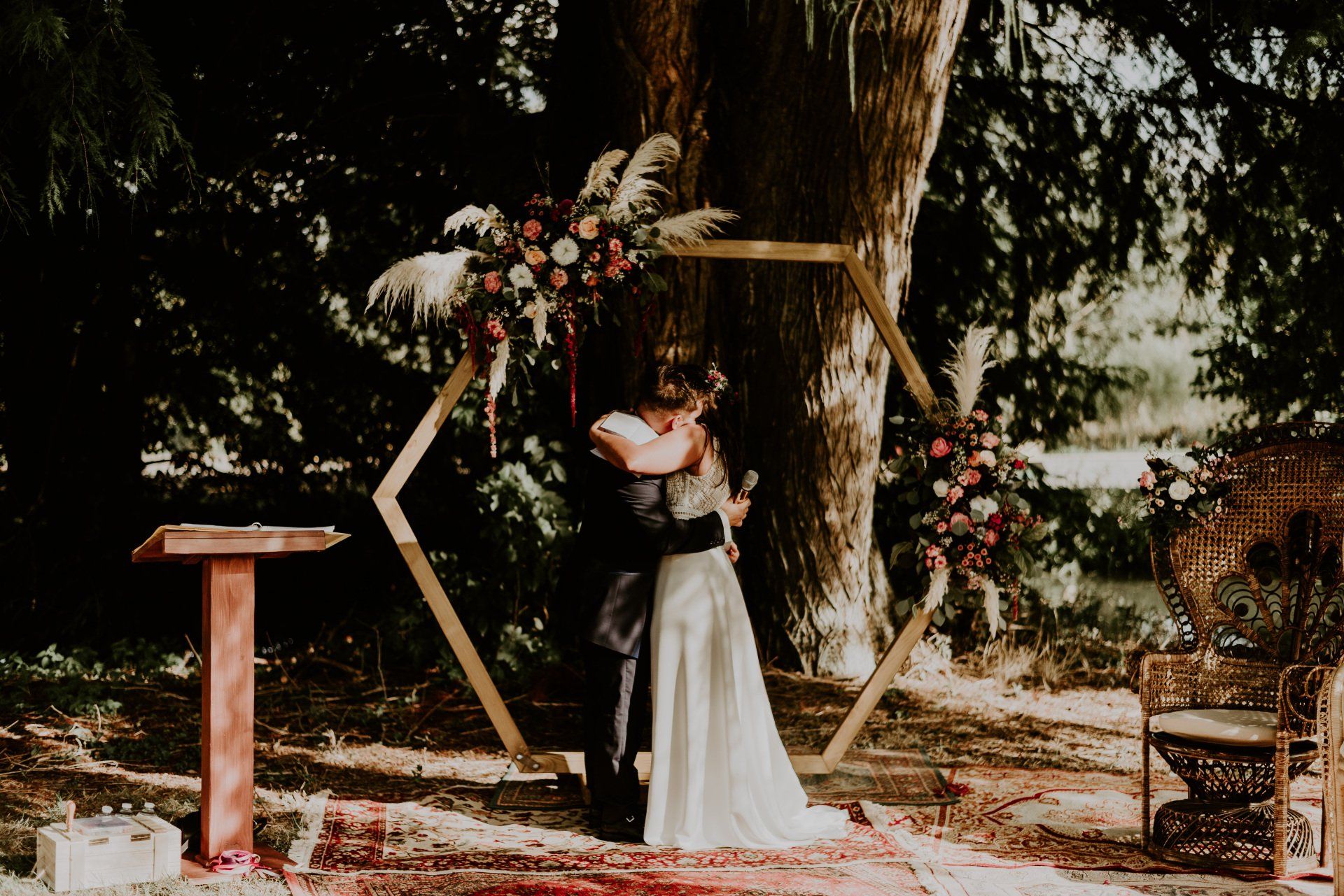 Cérémonie Laïque en forêt Photo d'une cérémonie laïque dans un parc arboré, deux mariés s'enlacent devant une arche hexagonale en bois, fleuries, sur des tapis persans, à côté de fauteuils emmanuelle en osier , fleuris