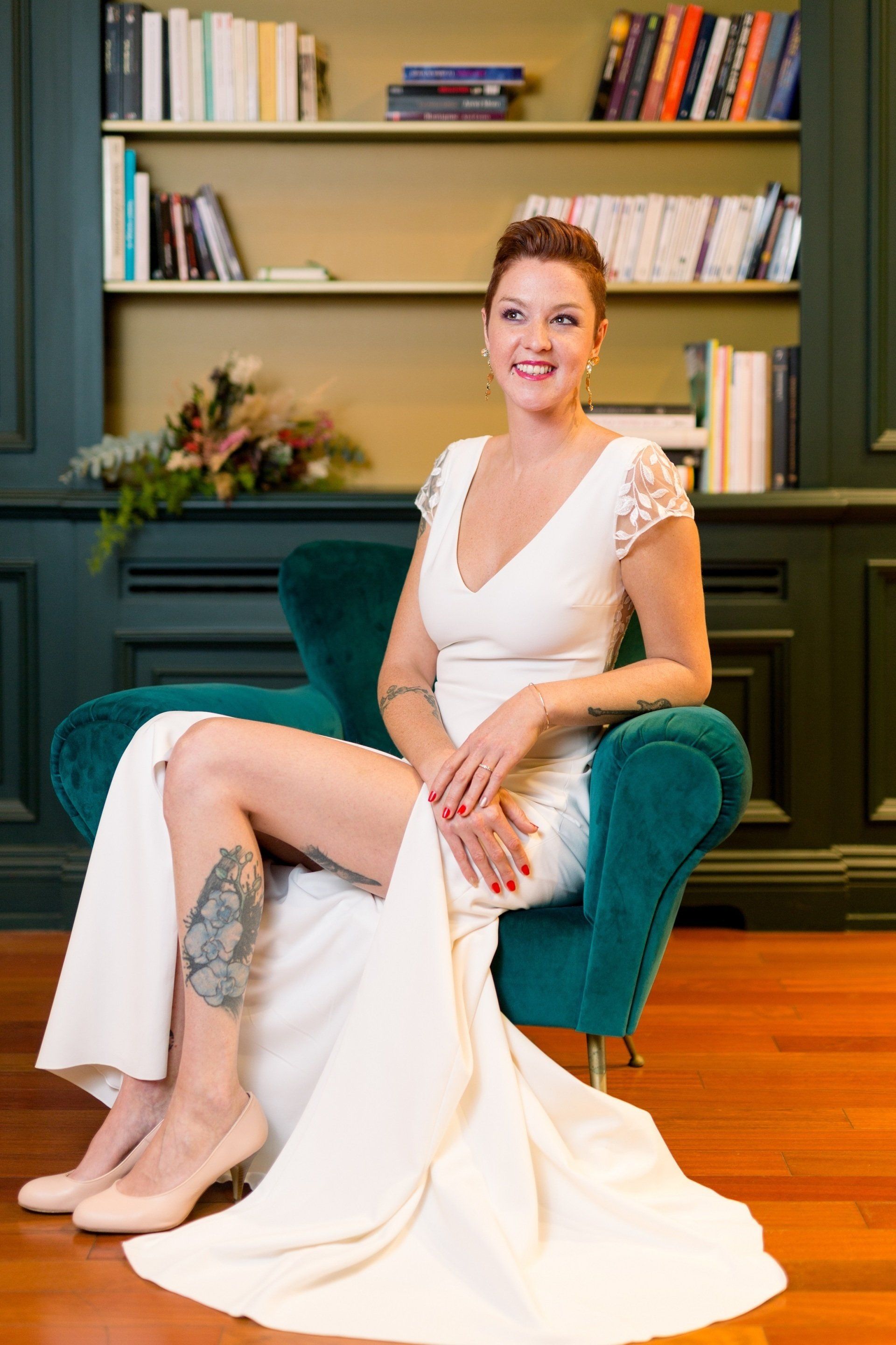 Mannequin femme , assise sur un fauteuil de velours bleu canard, dans un salon avec parquet ancien, devant une bibliothèque. Elle portant une robe de mariée ALICE SPRINGS MARIAGE , avec une fente au niveau des jambes. Robe à manches courtes dentelles. - Organisation Emi Wedding - Pays Basque