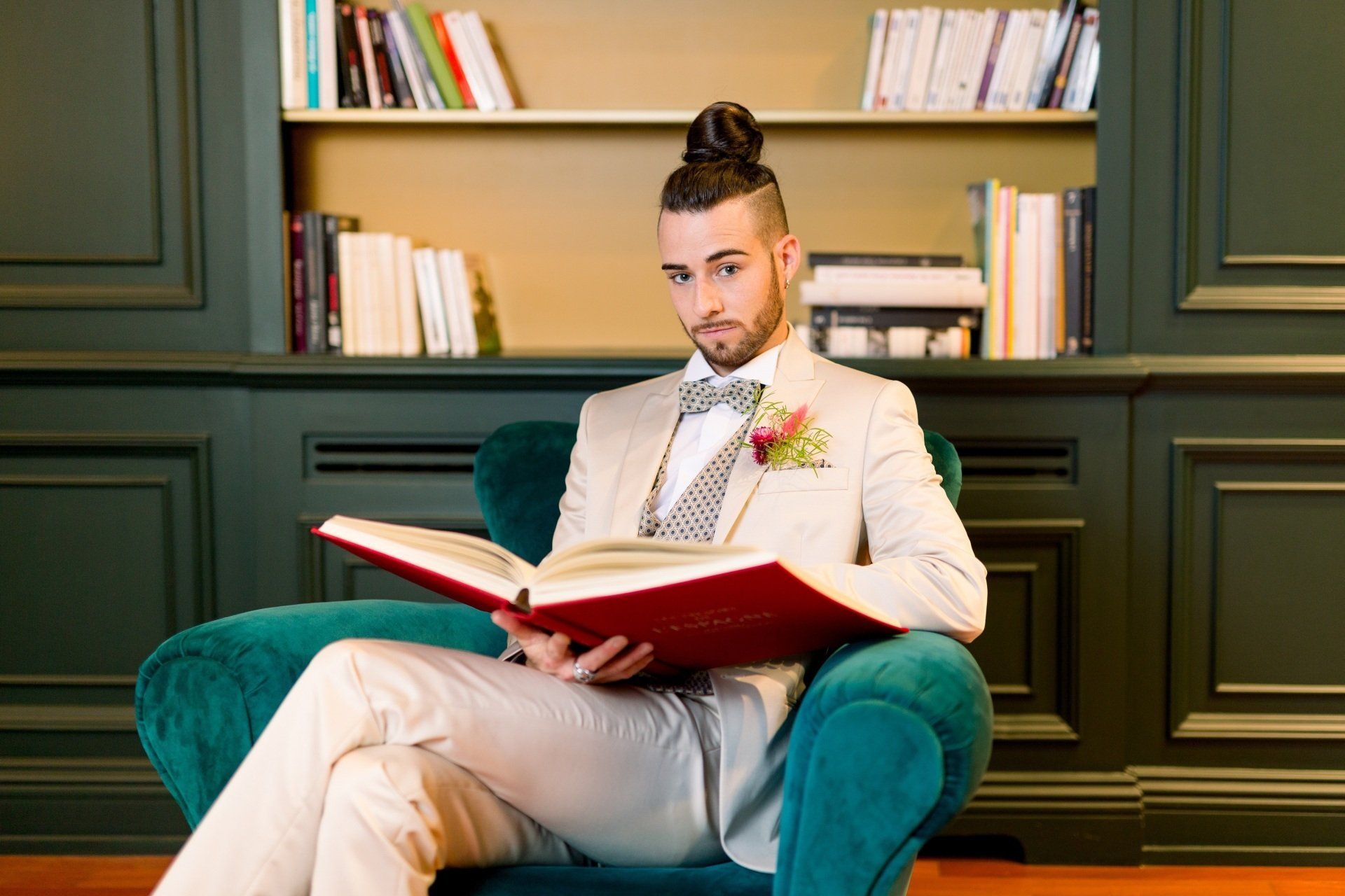 Mannequin homme assis sur un fauteuil de velours bleu canard, avec un livre ancien à couverture rouge ouvert sur les genoux, en costume de marié trois pièces beige avec boutonnière fleurie - Mise en scène Emi Wedding Pays Basque