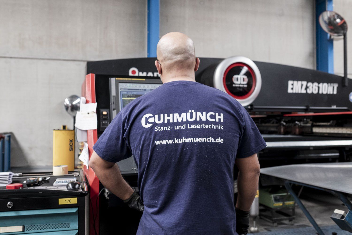 Ein Mann bedient, uns mit dem Rücken zugewandt eine Maschine. Auf seinem T-Shirt steht: Kuhmünch. Schweiß und Lasertechnik. Sowie der Name der Webseite.