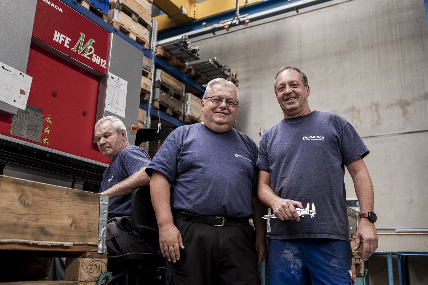 Zwei handwerkliche Männer lächeln in der Werkstatt in die Kamera, während ein dritter Mann seiner Arbeit nachgeht.