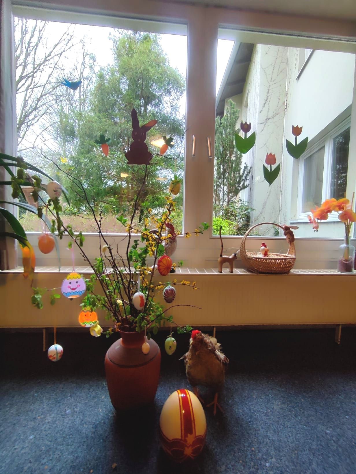 Osterdekoration auf einer Fensterbank mit geschmücktem Osterstrauß, bunten Ostereiern und Frühlingsblumen vor einem Fenster.