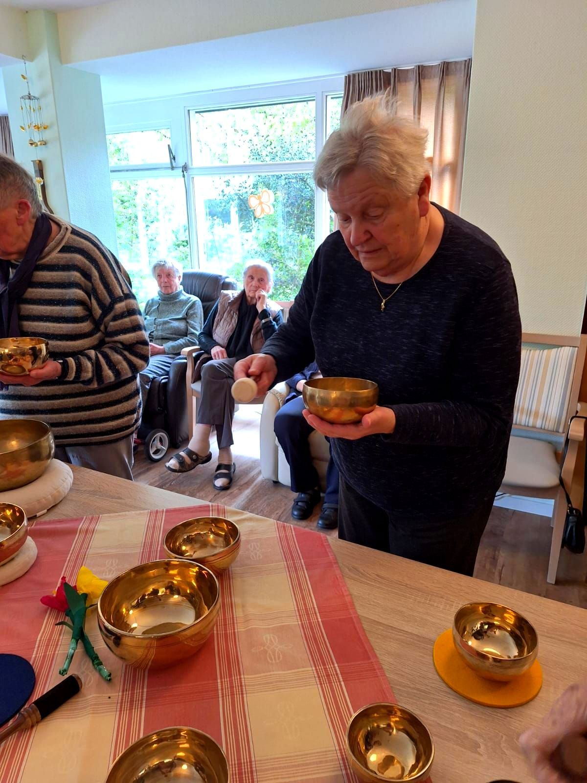 Bewohnerin schlägt eine Klangschale, mehrere Klangschalen stehen auf dem Tisch und andere Bewohnerinnen schauen zu.
