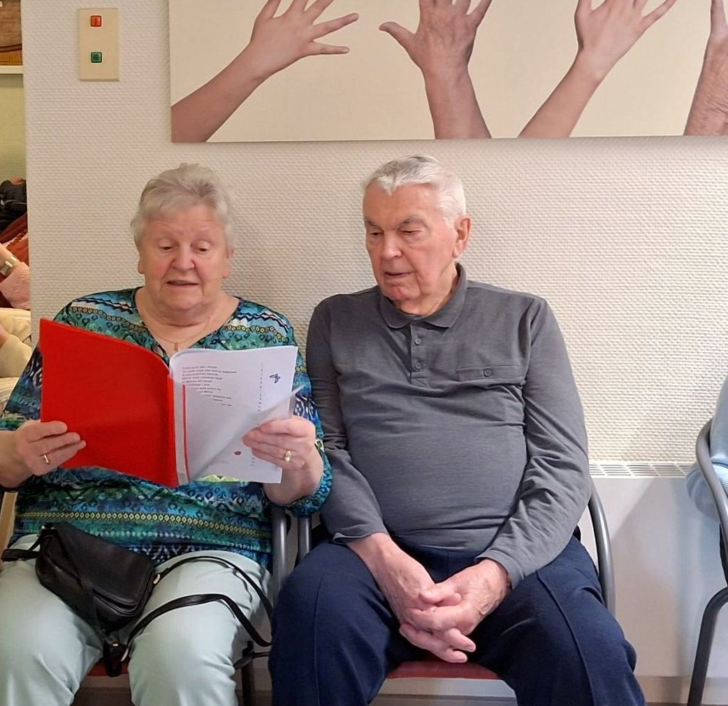 Bewohner und Bewohnerin des Lambertistifts sitzen nebeneinander und Singen mit einer roten Mappe in der Hand.