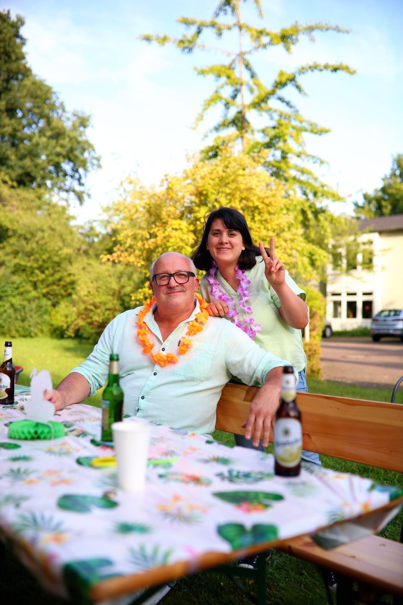 Mitarbeiter sitzt am Tisch beim Sommerfest im Garten, eine Kollegin steht hinter ihm und zeigt das Peace-Zeichen.