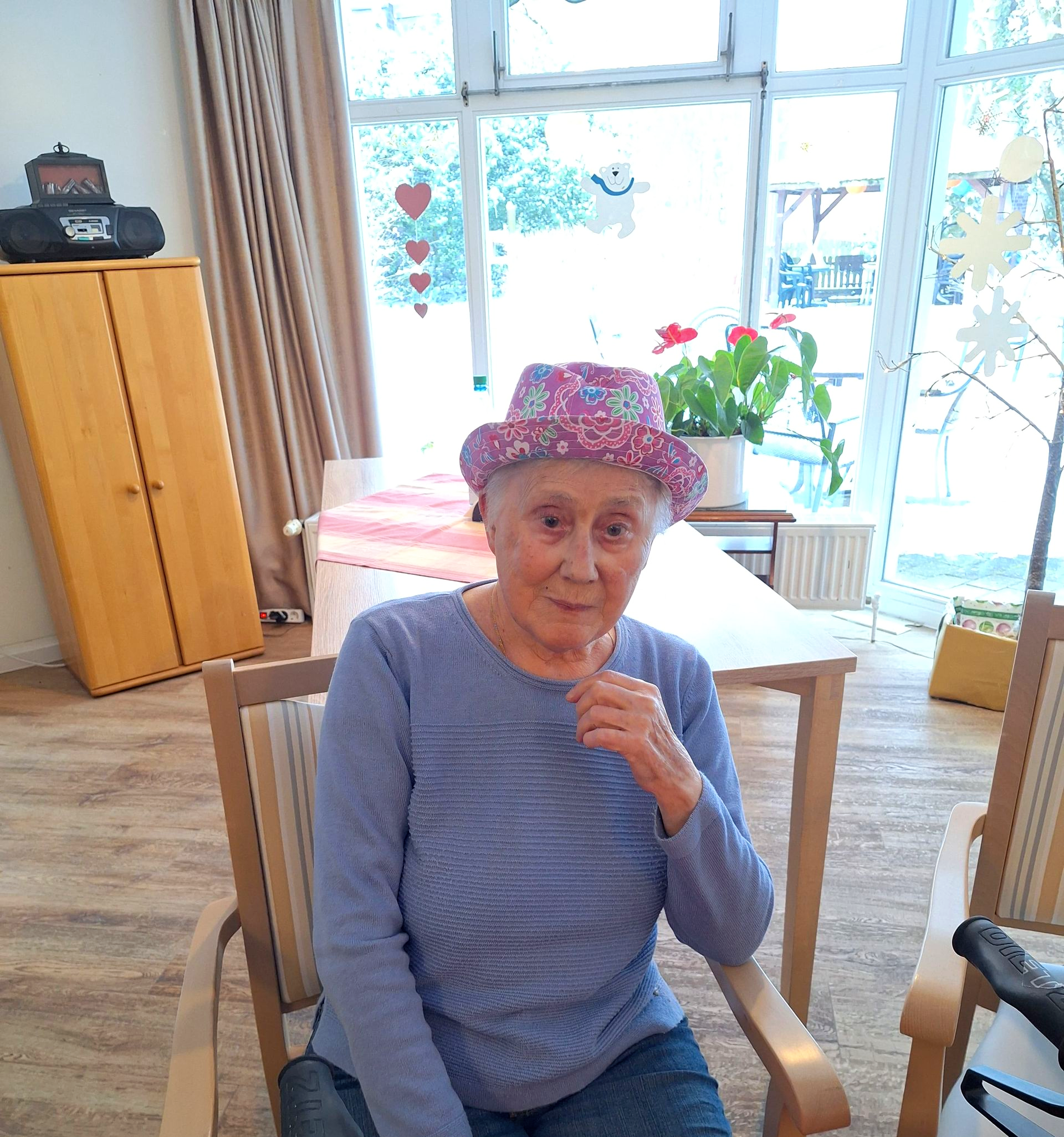 Eine Bewohnerin mit buntem Hut sitzt an einem Tisch im Aufenthaltsraum.