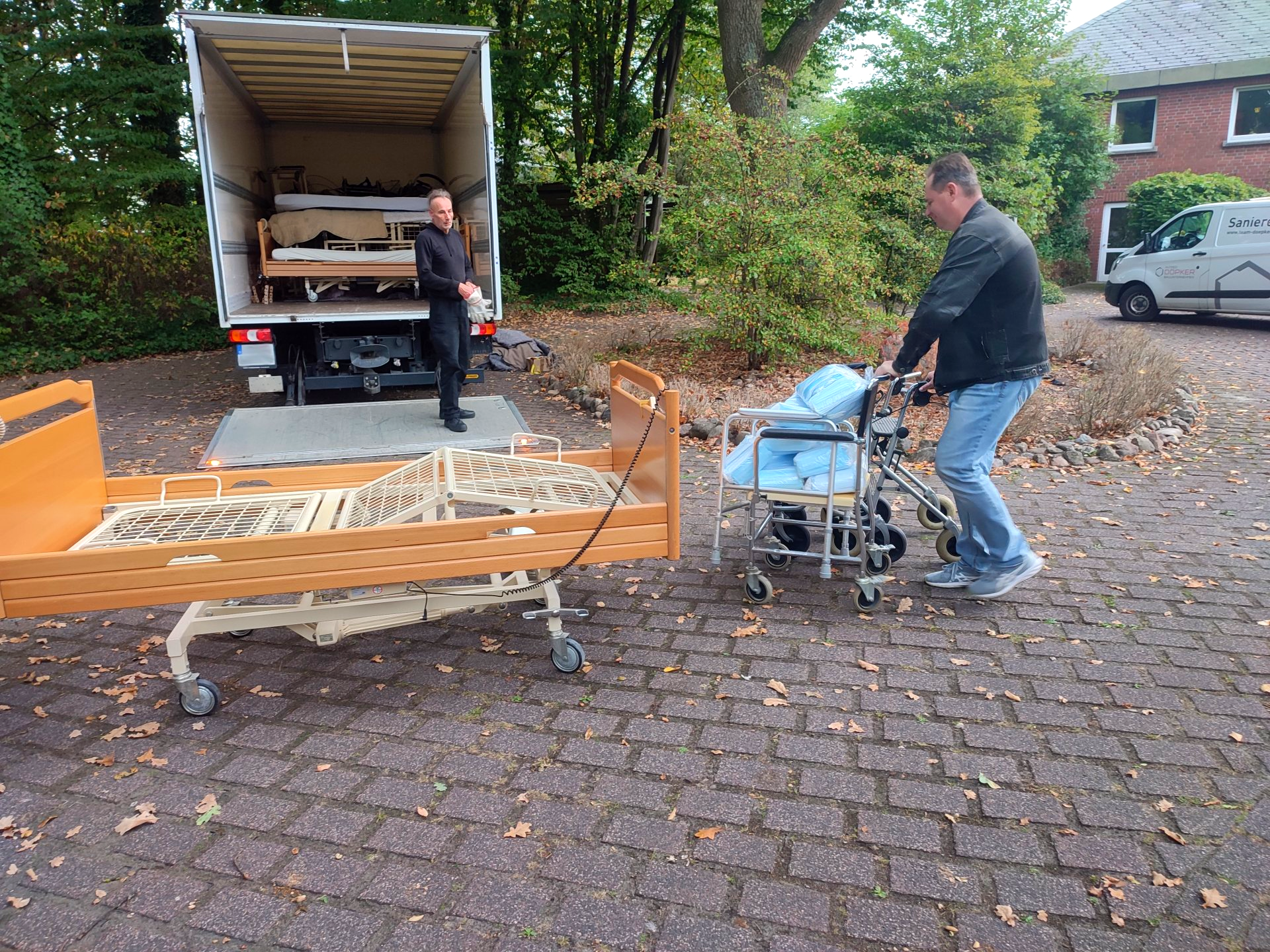 Person schiebt einen Rollstuhl mit Pflegehilfsmitteln zu einem Transporter.