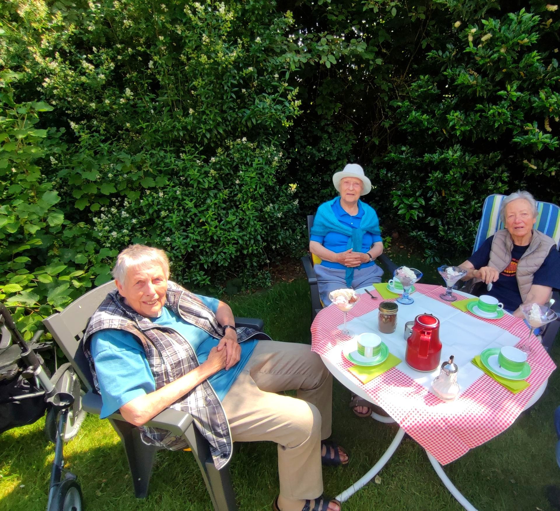 Drei Bewohnerinnen sitzen im Garten an einem Tisch und genießen gemeinsam Eis und Kaffee.