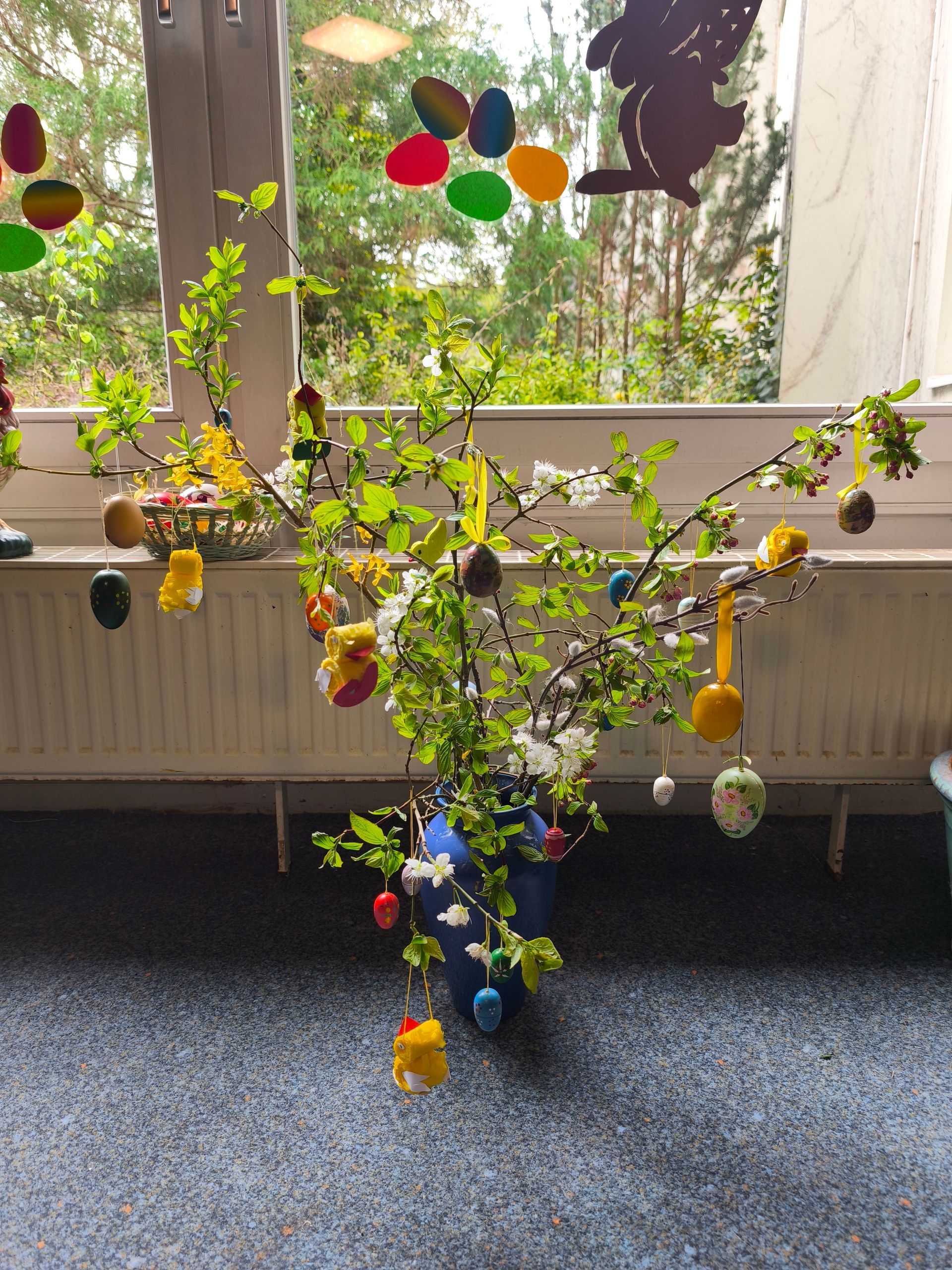 Ein geschmückter Osterstrauß mit bunten Ostereiern steht vor einem Fenster.