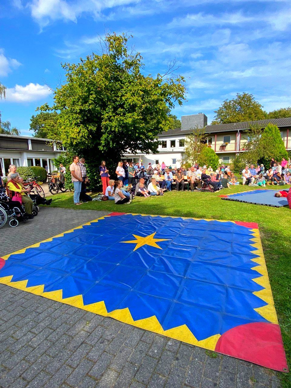 Viele Bewohner und Gäste sitzen im Garten und schauen einer Zirkusaufführung auf einer großen Matte zu.