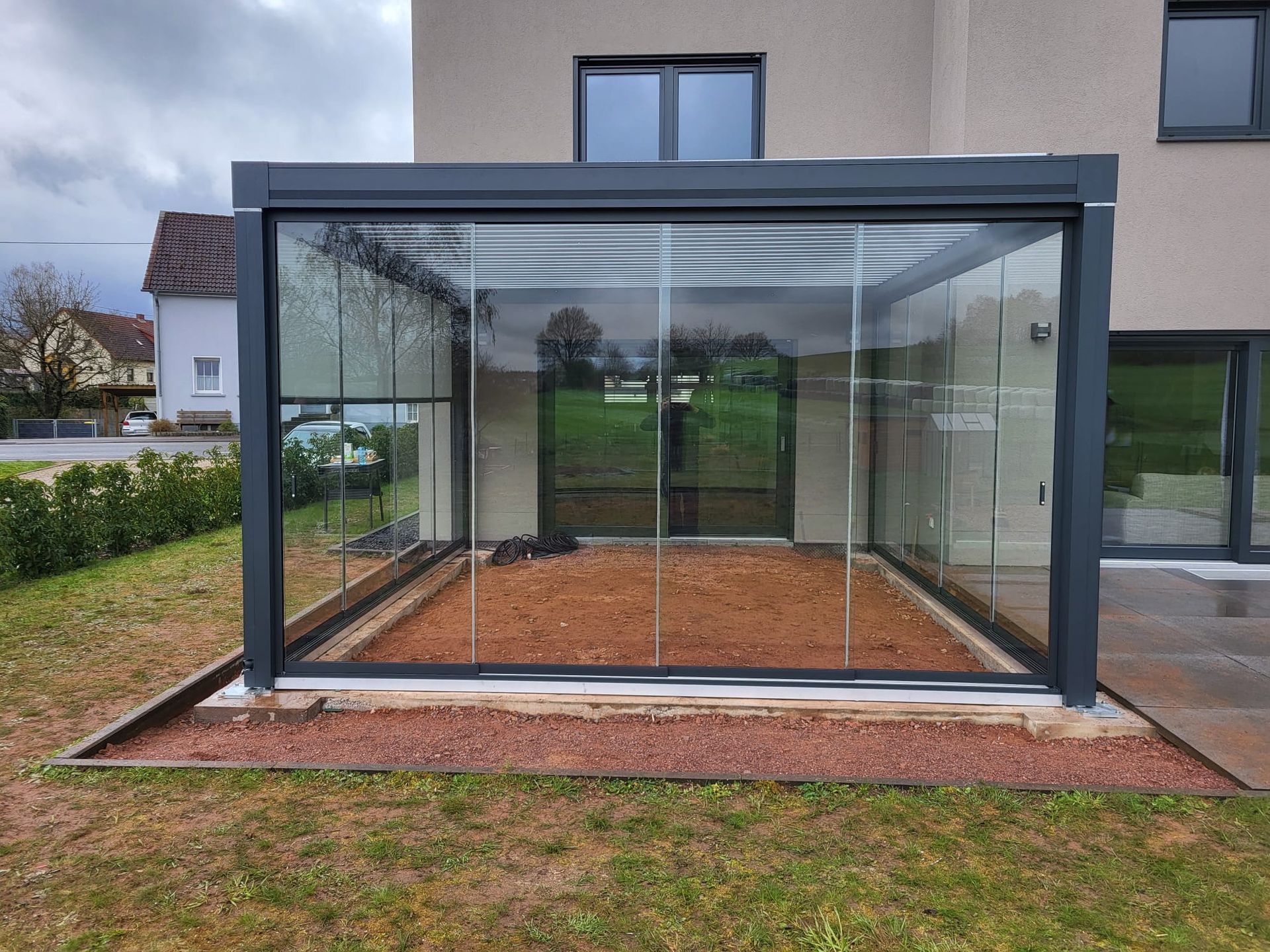 Lamellendach_weinor_neu_Saarland_Rolladen_Kessler modernes Haus mit Flachdach, davor auf das Streifenfundament ein Lamellendach in anthrazit gebaut, das Lamellendach ist unten mit Ganzglas-Schiebewänden geschlossen. Jetzt kann der Bodenbelag gelegt werden