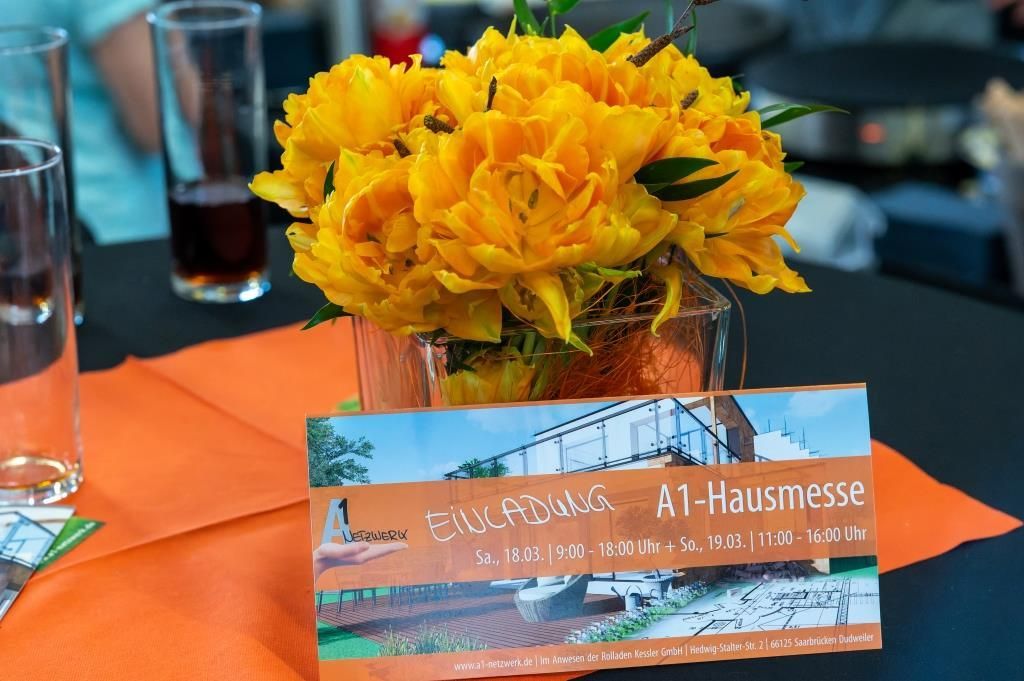 Eine Einladungskarte zur A1-Hausmesse auf einem orangenen Stehtisch mit orangenen Blumen. 