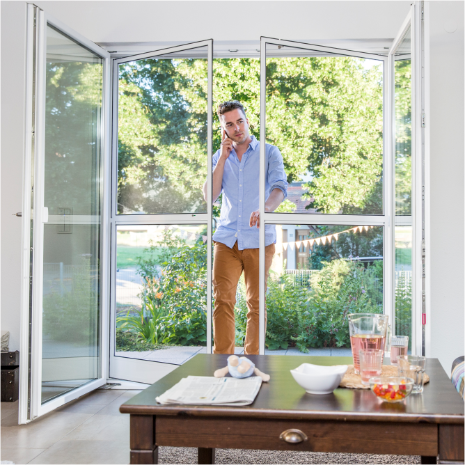 Mann geht durch eine zweiflüglige Insektenschutztür und tritt in sein Wohnzimmer. Draußen scheint die Sonne.