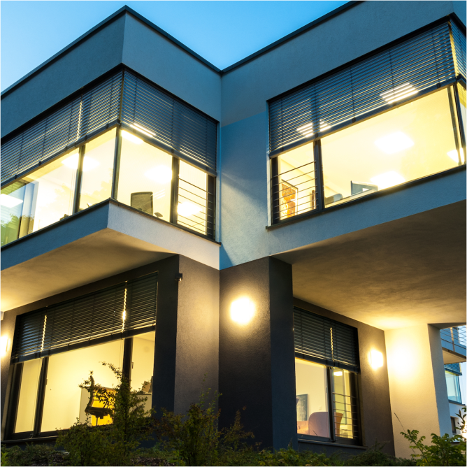 Ein kubisches Haus mit großen Glasfronten im Erd- und Obergeschoss. Es ist Dämmerung und drinnen ist das Licht an. Im Erdgeschoss sind Rollläden und im Obergeschoss Jalousien bzw. sogenannte Raffstores halb heruntergelassen.