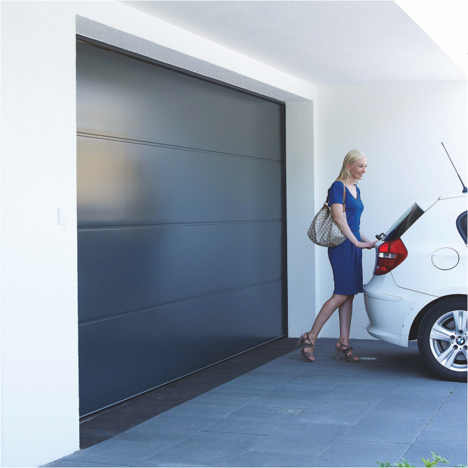 Ein geschlossenes Sectionaltor von Teckentrup mit glatter Oberfläche in Anthrazitgrau. Vor dem Tor steht eine Frau am Kofferraum ihres Autos, bereit für den Tag. Die klare, glatte Oberfläche des Tores verleiht dem Bild eine moderne und ansprechende Ästhetik.