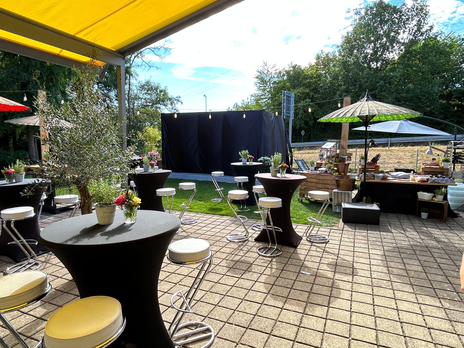 Im Garten unseres Anwesens in Saarbrücken sind bei sonnigen Wetter Stehtische, Sonnenschirme und Essenstände im spanischen Flair aufgebaut. Das Lamellendach Artares ist noch in schwarze Tücher gehüllt.