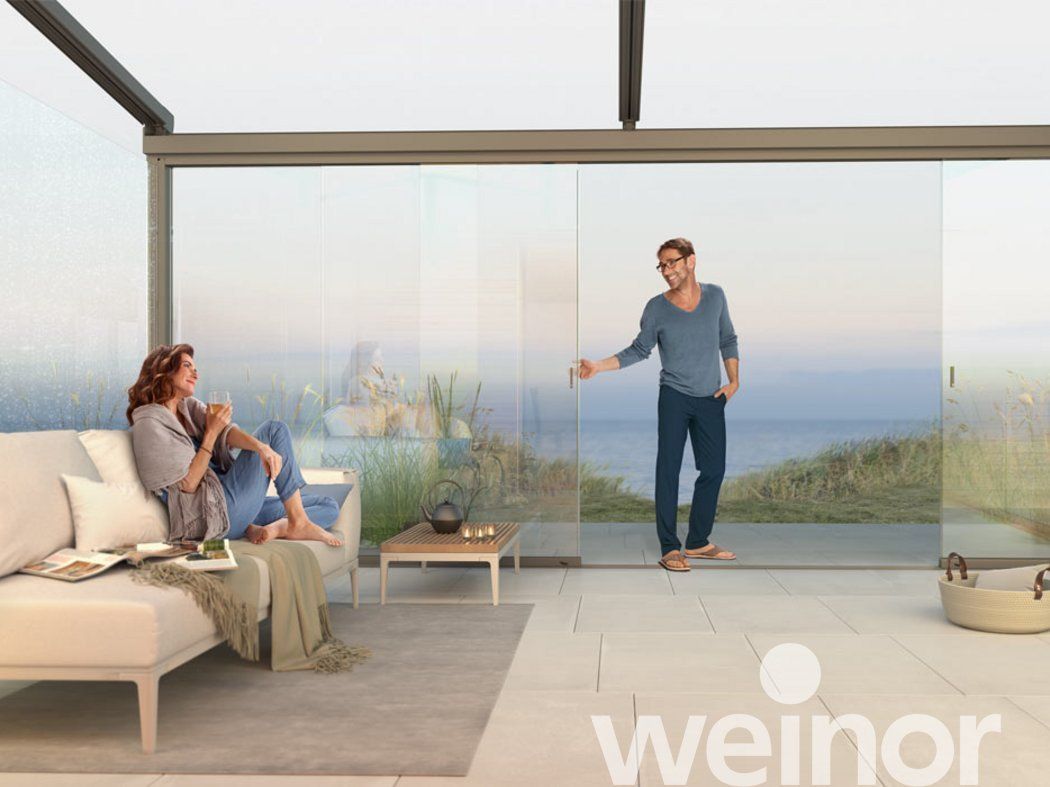 Blick von Innen unter einem Lamellendach von weinor. Eine Frau sitzt auf den Gartenmöbeln, der Mann zieht locker mit einer Hand die Ganzglasschiebetür von weinor zu.