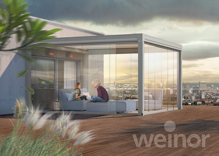 Blick auf eine Holzterrasse über der Stadt. Darauf steht ein Lamellendach von weinor, welches mit Ganzglasschiebetüren rundherum geschlossen ist.