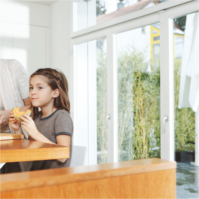 Ein Mädchen sitzt am Frühstückstisch vor einer beeindruckenden Fensterfassade. Sie genießt ihr Frühstück, während das Tageslicht durch die großen Fenster einfällt und eine angenehme Atmosphäre schafft.