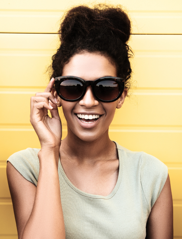 Junge Frau trägt große dunkle Sonnenbrille, lächelt dabei.