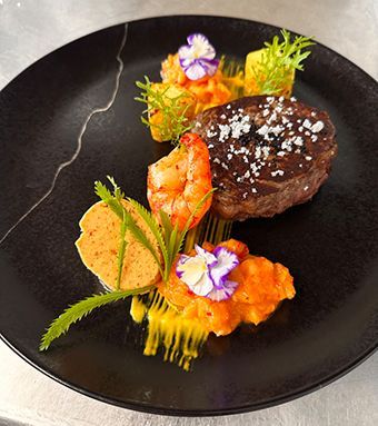 Elegant angerichtetes Hauptgericht mit Fleisch, Garnelen und essbaren Blüten auf schwarzem Teller – Schlosscafé Teningen