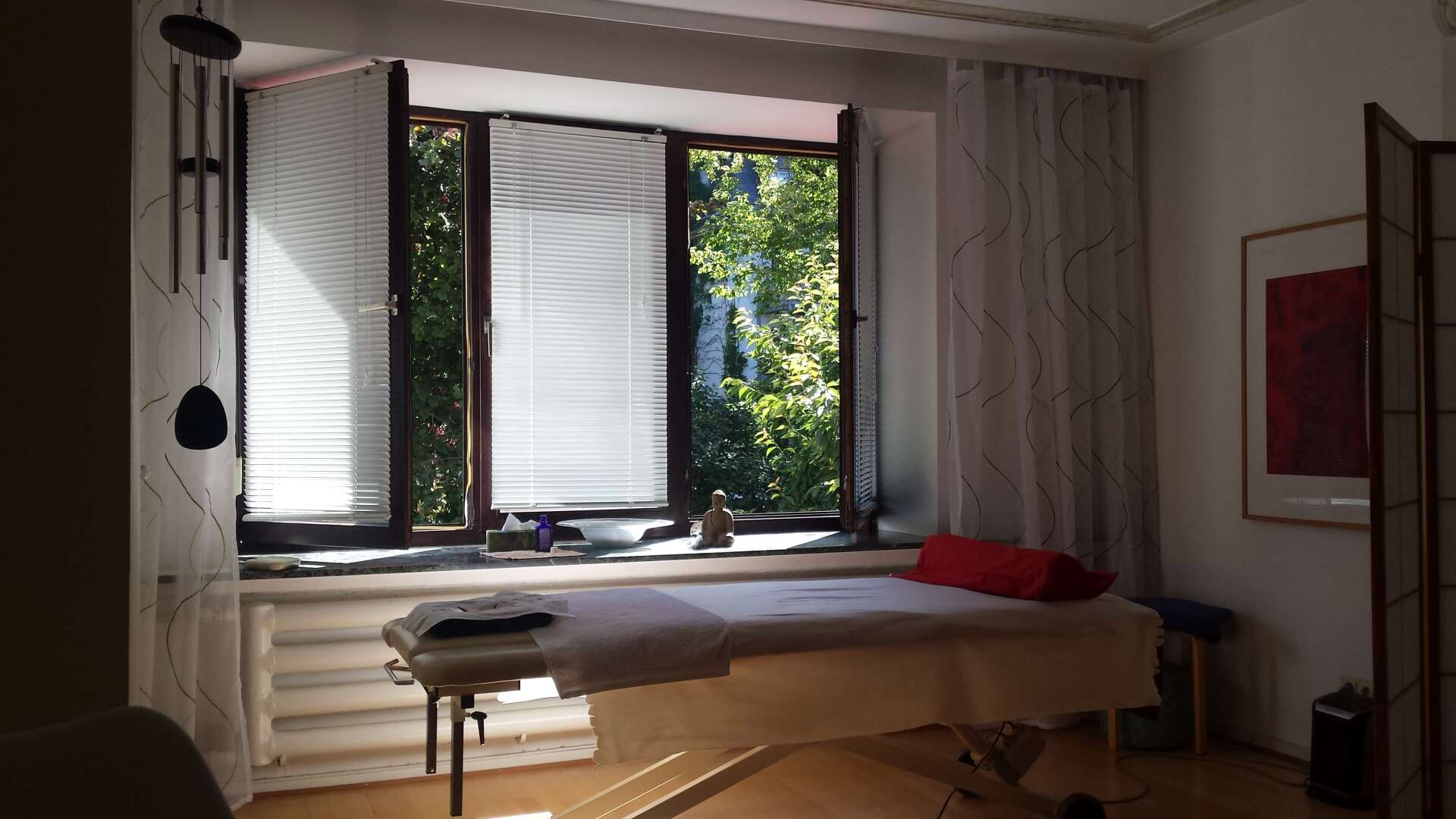 Entspannungsmassagen Köln Massageliege vorgeöffnetem Fenster im abgedunkelten Raum mit Blick in den Garten