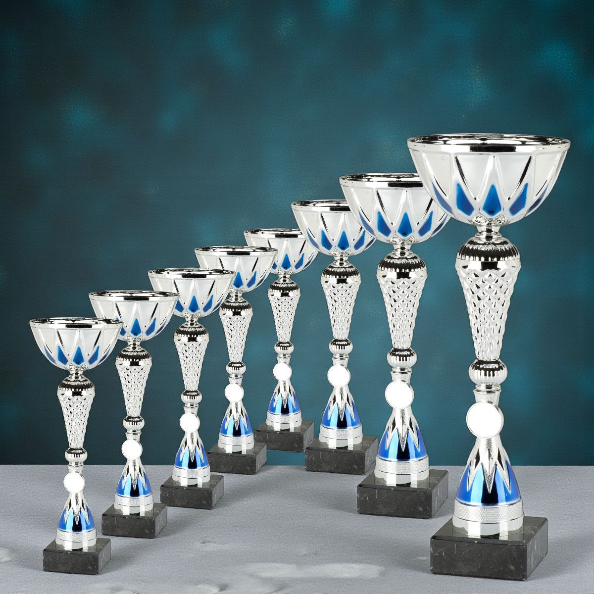 „Gold-, Silber- und Bronzepokal auf Marmorsockeln vor hellgrau-blauem Hintergrund.“