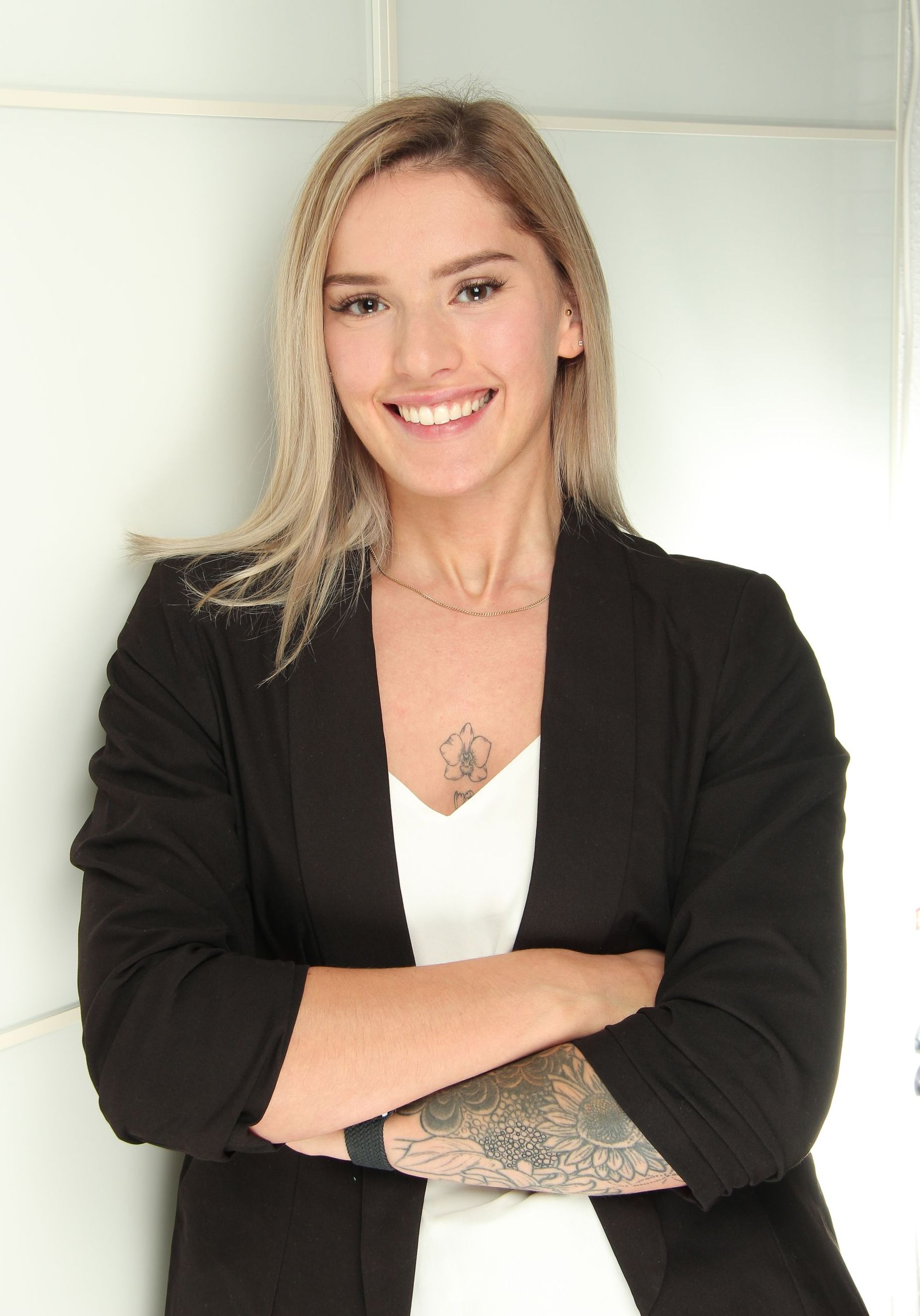 Das Bild zeigt ein Businessportrait einer Jungen, blonden Frau mit verschränkten Armen. Sie lächelt freundlich und offen. Sie trägt einen schwarzen Blazer, darunter ein weißes Oberteil mit V-Ausschnitt auf dem Dekolletee sieht man ein Blumentatto. Der linke Unterarm ist ebenfalls mit Blumen tätowiert. Sie steht vor einer leicht grünlichen Glaswand.