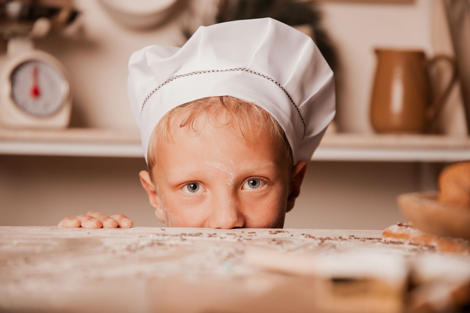 Weihnachtsbäckerei Ein kleiner junge guckt über einen, mit Mehl bedeckten Tisch. Der Mund ist hinter dem Tisch, Nase und Augen sind zu sehen und das Gesicht ist ein wenig mit Mehl bedeckt. Die Finger einer Hand liegen auf dem Tisch. Der Junge hat Grüne Augen, blondes Haar und Augenbrauen und eine weiße Kochmütze auf. Im Hintergrund sind ein brauner Krug und eine weiße Küchenwage unscharf zu erkennen.