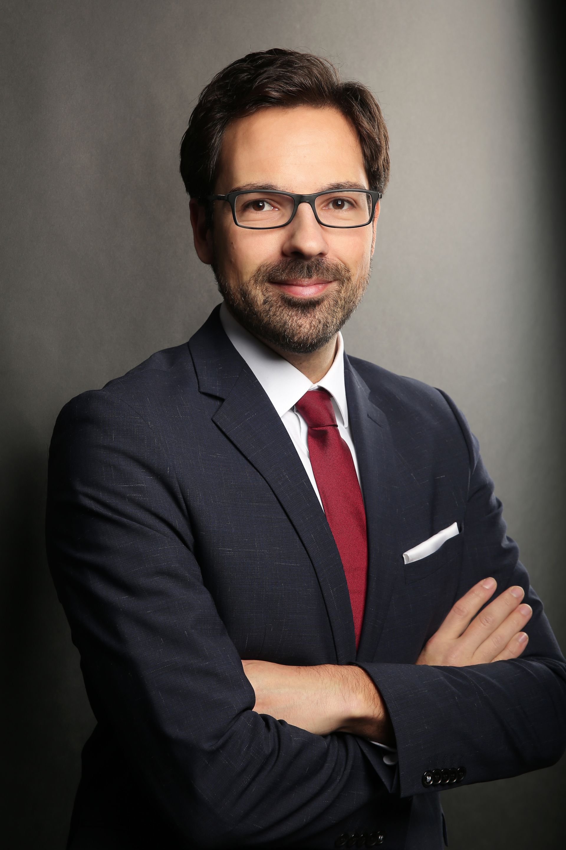 Businessportrait eines jungen Mannes mit verschränkten Armen vor dunkelgrauem, beleuchtetem Hintergrund. Der Mann hat Dunkle, etwas längere Haare und einen dunklen Kinn- und Oberlippenbart. Er trägt eine eckige Brille mit dunklem Gestell, ein dunkelblaues Sakko mit einem weißen Einstecktuch, ein Weißes Hemd und eine rote Krawatte. Der Mann lächelt mit geschlossenem Mund und hat braune Augen.
