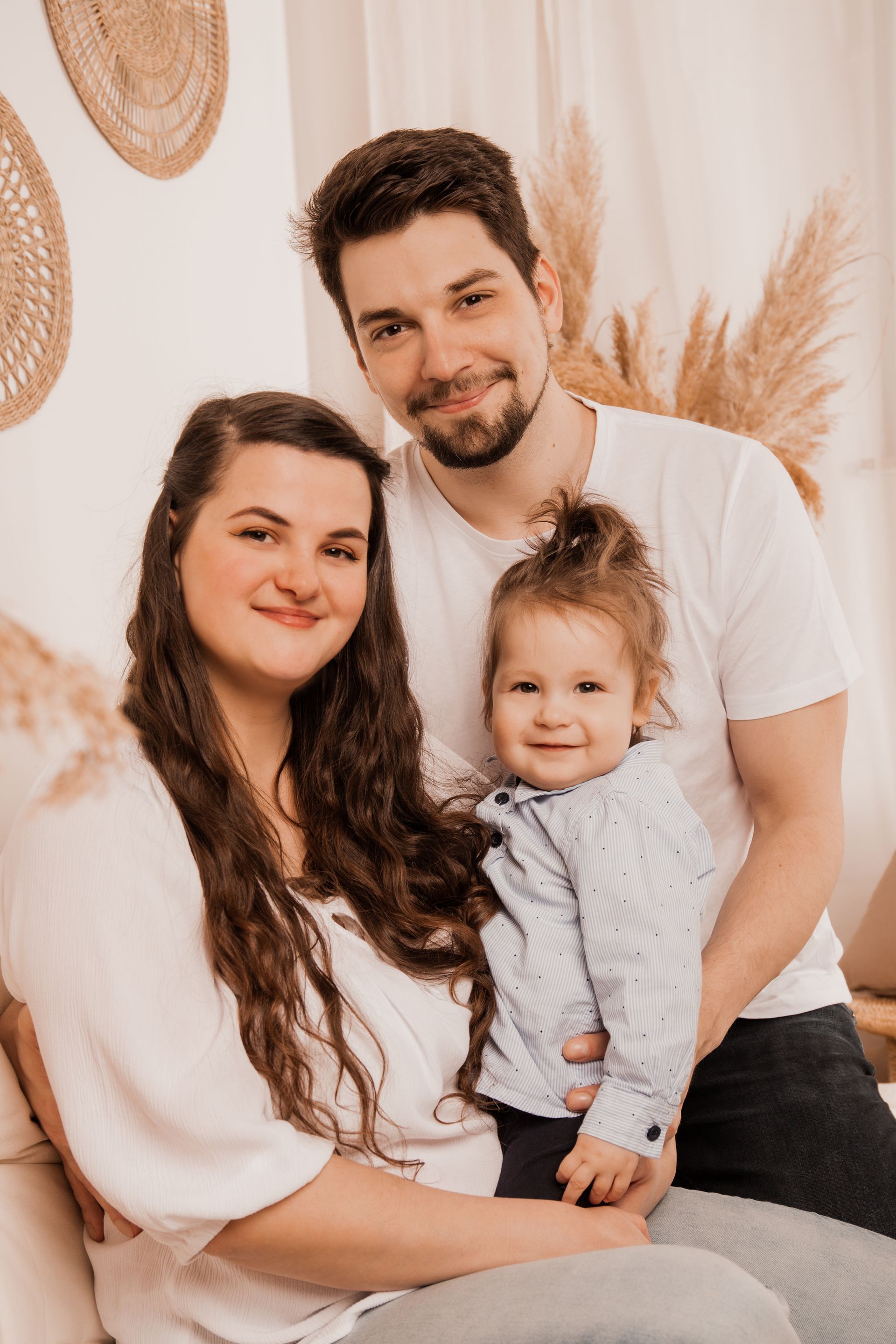Das Bild zeigt eine junge Familie: Mama, Papa und Sohn. Die Mama hat eine weiße Bluse an und lange, braune Haare. Der Papa hat ebenfalls braune Haare (etwas länger) und einen braunen kurzenOberlippen- und Kinnbart und trägt ein weißes T-shirt. Der Sohn hat lange braune Haare zu einem hohen Zopf gebunden. Er trägt ein hellblaues Hemd mit blauen Punkten. Alle lächeln mit geschlossenem Mund und schauen in die Kamera. Das Bild zeigt nur Oberkörperausschnitte, ist in einem warm-braunen Stil. Im Hintergrund ist ein wenig Pampasgras, sowie teile von Rattan Elementen zu sehen.